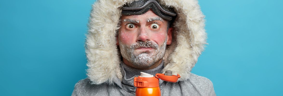 Puzzled frozen man in winter clothes tries to warm himself with hot beverage has red face and bear covered with blizzard spends much time outdoors during snowboarding. Frosty weather conditions